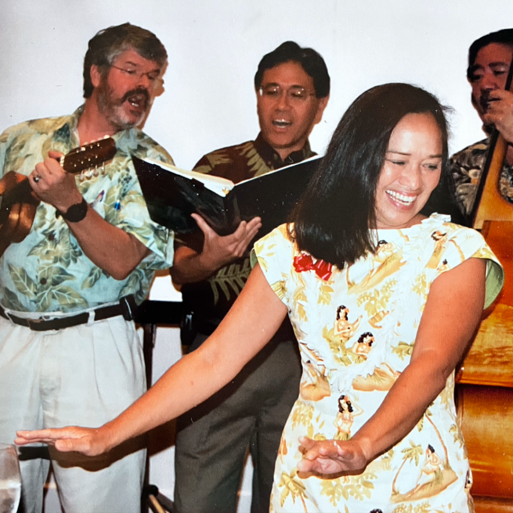 Mele Look dancing hula with 3 musicians in the background, singing